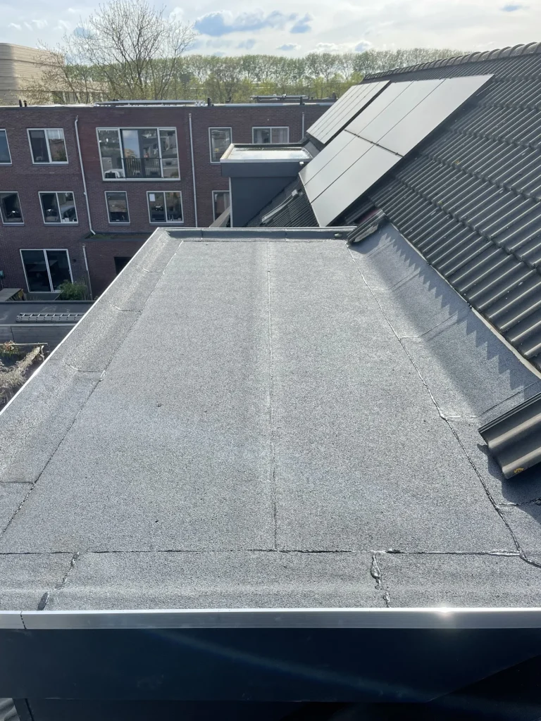 Nieuwe bitumen dakbedekking met aluminium daktrim op een aanbouw naast een pannendak met zonnepanelen